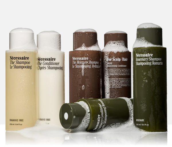Six Nécessaire haircare bottles arranged on a white studio background, including shampoo, conditioner, and scalp hair products. The bottles are in cream, white, brown, and olive green, with some foam visible at the top and one bottle lying on its side in front.