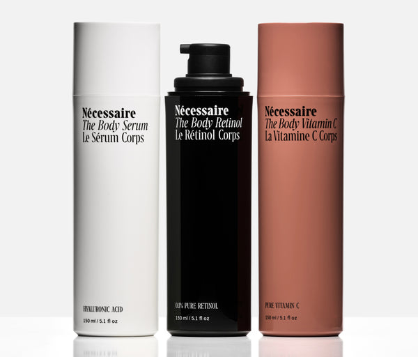 Three cylindrical skincare bottles from Nécessaire on a light gray background: a white bottle labeled The Body Serum - Hyaluronic Acid, a black bottle labeled The Body Retinol - 100% Pure Retinol, and a terracotta-colored bottle labeled The Body Vitamin C - Pure Vitamin C.